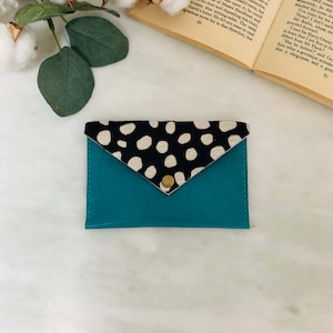 May include: A teal leather cardholder with a flap closure. The flap is made of black and white spotted cowhide and secured with a gold snap. The cardholder is a stylish accessory for carrying credit cards and IDs.