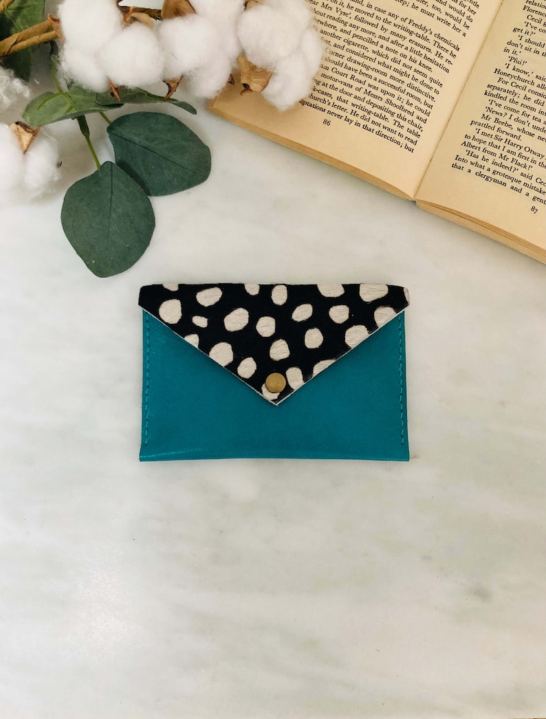May include: A teal leather cardholder with a flap closure. The flap is made of black and white spotted cowhide and secured with a gold snap. The cardholder is a stylish accessory for carrying credit cards and IDs.