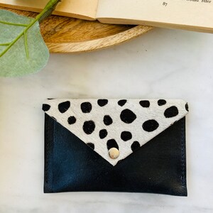 May include: Black leather card holder with a flap closure. The flap is made of cowhide with black spots on a white background. A silver snap secures the flap. The card holder is on a white surface.