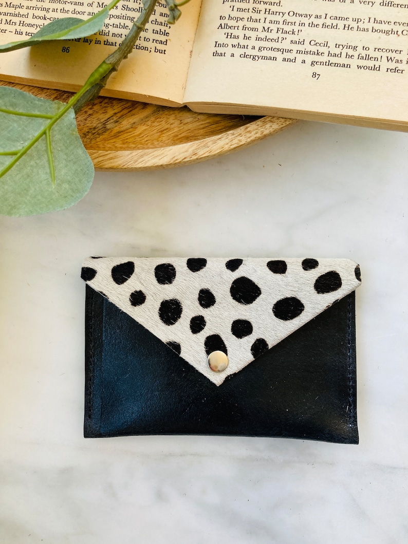 May include: Black leather card holder with a flap closure. The flap is made of cowhide with black spots on a white background. A silver snap secures the flap. The card holder is on a white surface.