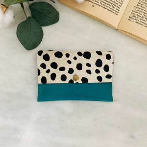 May include: A small, rectangular wallet with a teal bottom and a top section featuring a black and white spotted pattern. The wallet is secured with a gold-colored snap closure. The wallet is likely made of leather.
