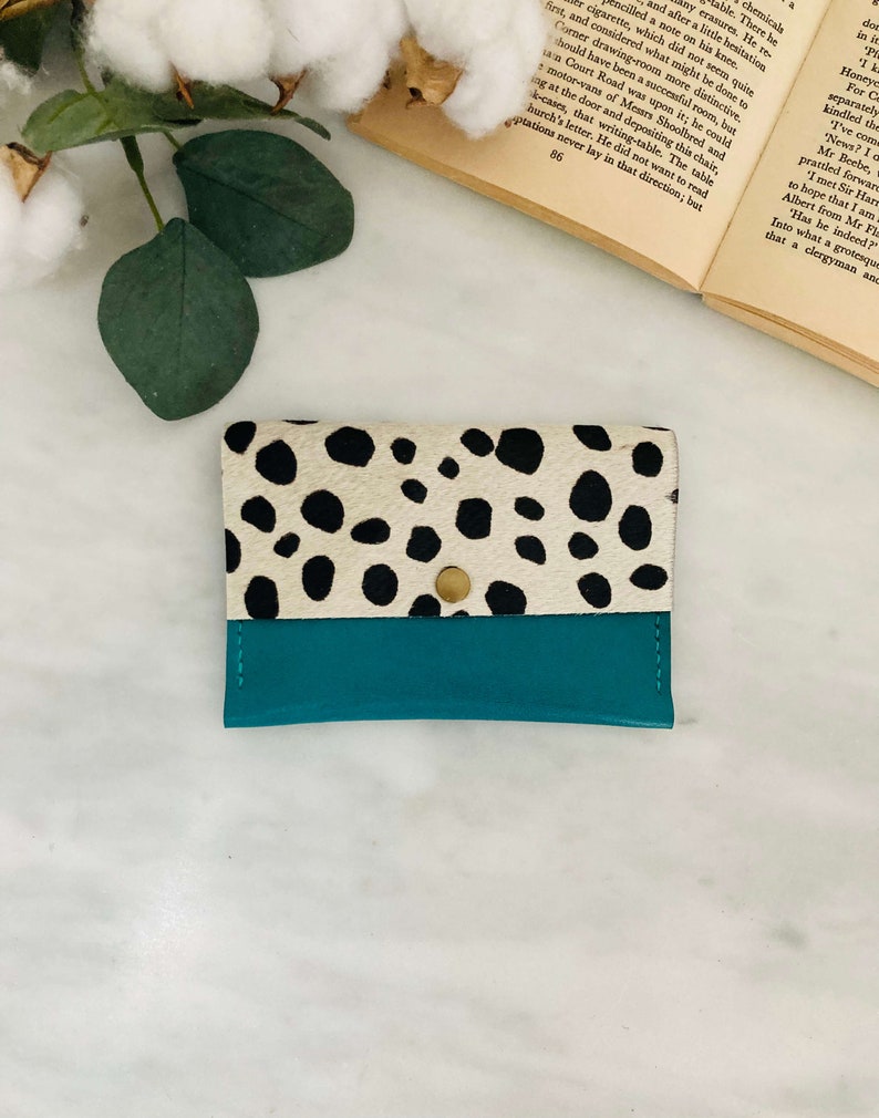 May include: A small, rectangular wallet with a teal bottom and a top section featuring a black and white spotted pattern. The wallet is secured with a gold-colored snap closure. The wallet is likely made of leather.