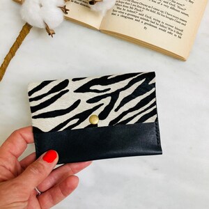 May include: A black and white zebra print card holder with a black leather bottom flap and a gold snap closure. The card holder is made of genuine leather and is a stylish accessory.
