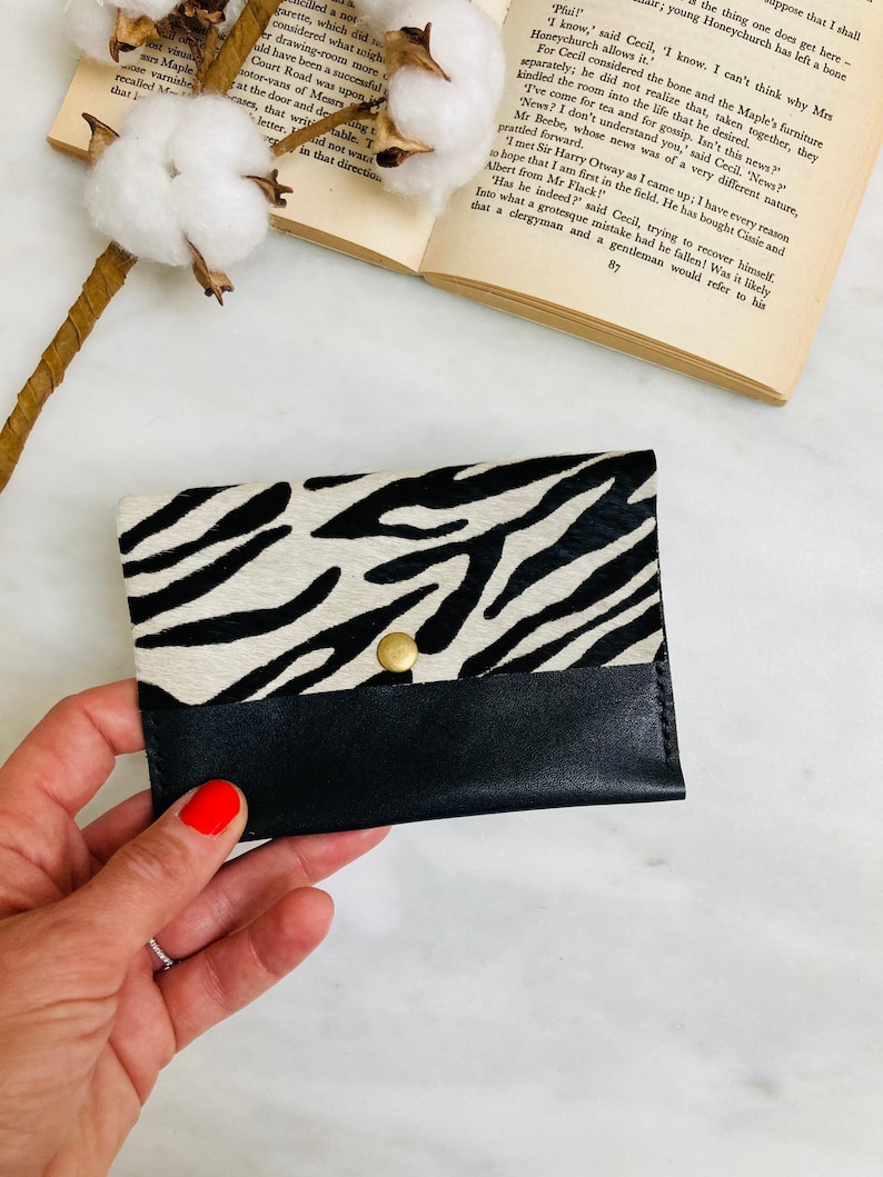 May include: A black and white zebra print card holder with a black leather bottom flap and a gold snap closure. The card holder is made of genuine leather and is a stylish accessory.