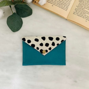 May include: Teal leather cardholder with a flap closure. The flap is made of cowhide with black spots. A gold-colored snap secures the flap. The cardholder is on a white surface with a book and greenery in the background.