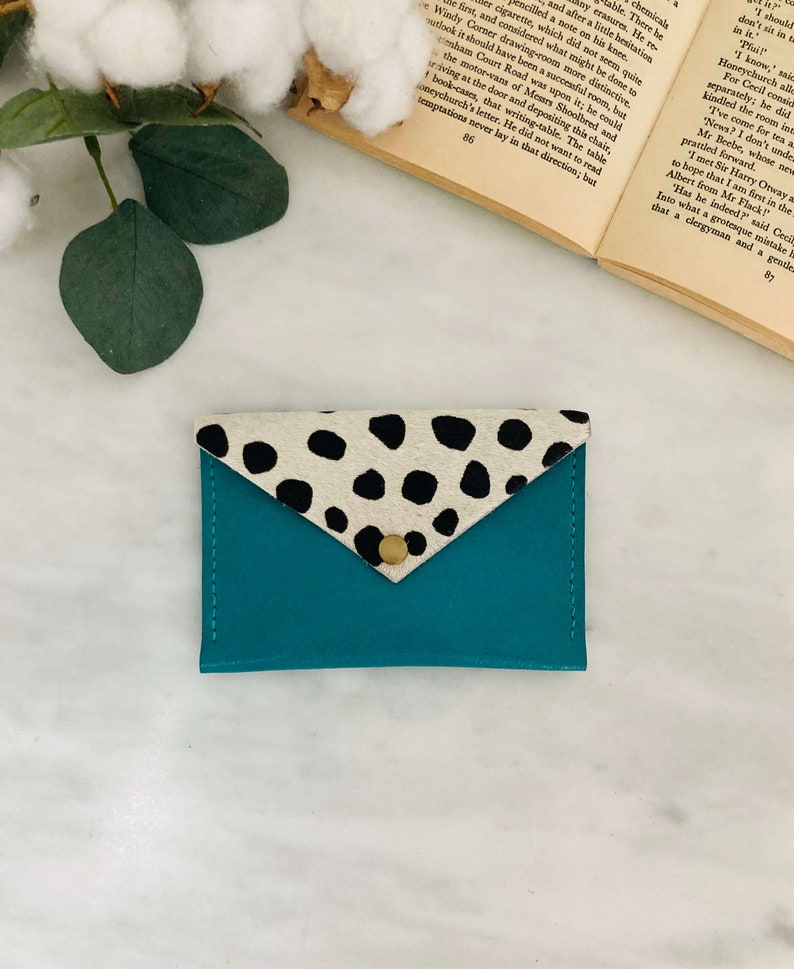 May include: Teal leather cardholder with a flap closure. The flap is made of cowhide with black spots. A gold-colored snap secures the flap. The cardholder is on a white surface with a book and greenery in the background.