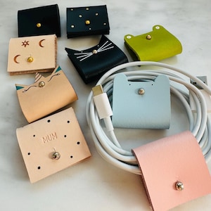 May include: A selection of leather cable tidies in various colours and designs. The organisers include black, light blue, pink, and beige options, some with decorative details like dots, stars, and cat faces. One organiser has the word "MUM" printed on it.