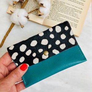 May include: A teal and black wallet with a cowhide pattern and a snap closure. The top portion of the wallet features a black and white spotted design, while the bottom is a solid teal color. The wallet is held by a hand.