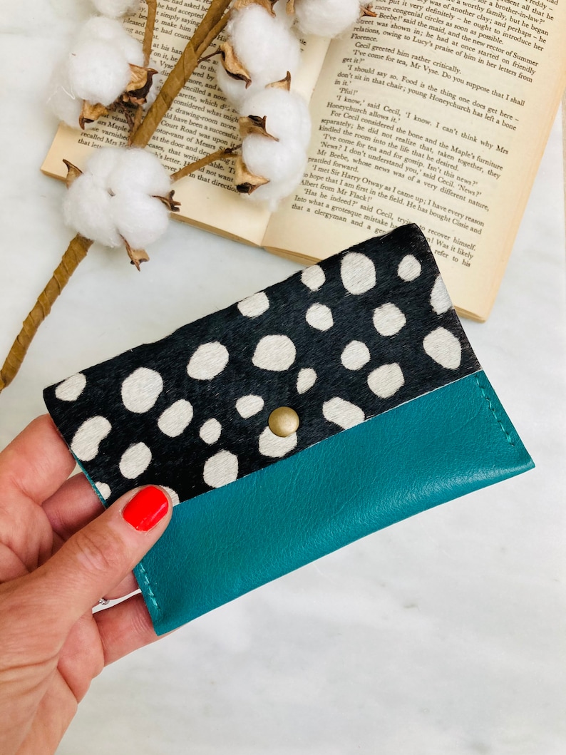 May include: A teal and black wallet with a cowhide pattern and a snap closure. The top portion of the wallet features a black and white spotted design, while the bottom is a solid teal color. The wallet is held by a hand.