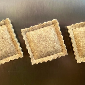 Op de afbeelding: Drie bruine ravioli-vormige kussens van vilt met een decoratieve gestikte rand.