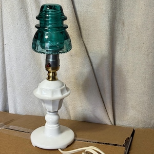 May include: A distinctive table lamp with a teal glass insulator top and a white ceramic base. The lamp has a silver and gold-toned metal socket and a white cord. The base is round with a decorative, fluted design. A vintage-inspired lighting fixture.