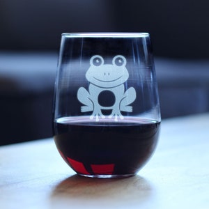 Puede incluir: Una copa de vino de vidrio transparente con un diseño de rana blanca grabado en el lateral. La copa está llena de vino tinto.