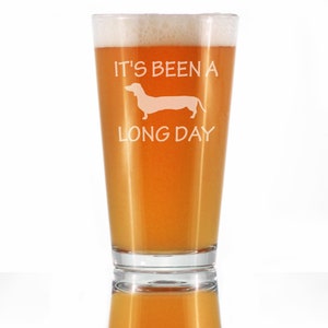 May include: A clear glass pint glass with a white dachshund silhouette and the text "It's been a long day". The glass is filled with a light amber colored beverage.