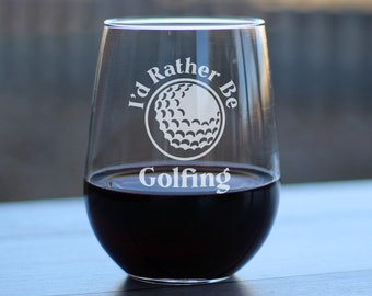 May include: A stemless wine glass filled with red liquid. The glass has the words "I'd Rather Be Golfing" etched in white, along with a golf ball graphic. The glass is clear, and the background is blurred.