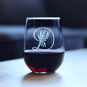Könnte beinhalten: Ein klares Weinglas aus Glas mit Stiel und einem weißen, geätzten Design eines stilisierten Buchstabens D mit einem Blattzweig im Inneren des Buchstabens. Das Glas ist mit Rotwein gefüllt.