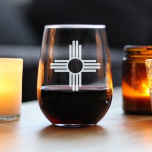 May include: A clear glass wine glass with a red wine inside. The glass has a white design of the Zia symbol, a traditional symbol of the Pueblo people of New Mexico.