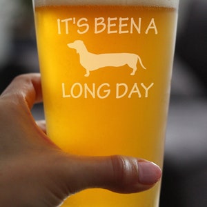 May include: A glass of beer with a white dachshund silhouette and the text "It's been a long day" on the side.