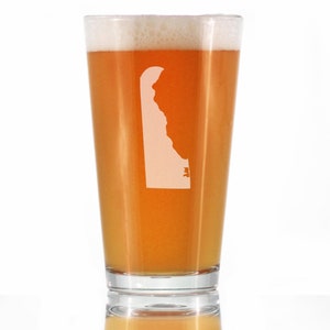 May include: A clear glass pint glass with a white outline of the state of Delaware on the side. The glass is filled with a light amber colored beer.