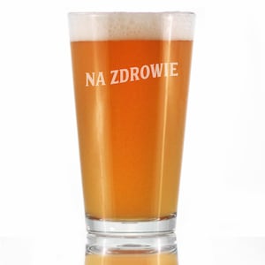 Puede incluir: Un vaso de cerveza alto de vidrio con una base de vidrio transparente y una parte superior de vidrio esmerilado. El vaso está lleno de una cerveza de color ámbar claro y tiene el texto "NA ZDROWIE" impreso en el lateral.
