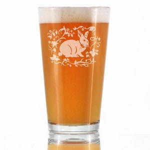 May include: A clear glass pint glass with a frosted design of a rabbit sitting in a floral wreath. The glass is filled with a light amber colored beer.