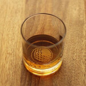 May include: A clear glass with a golf ball design on the bottom, filled with amber-colored liquid.