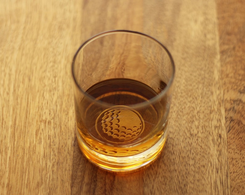 May include: A clear glass with a golf ball design on the bottom, filled with amber-colored liquid.