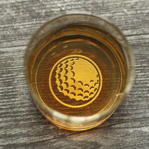 May include: A clear glass with a gold golf ball design on the bottom. The glass is filled with a golden liquid.