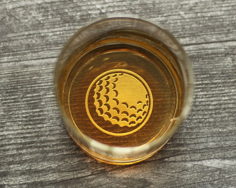 May include: A clear glass with a gold golf ball design on the bottom. The glass is filled with a golden liquid.