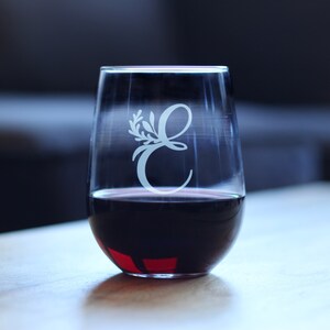 May include: A clear glass wine glass with a stem and a wide bowl. The glass is filled with red wine and has a white etched design of the letter "E" with a floral motif.