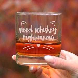 May include: A clear glass whiskey tumbler with the text "need whiskey right meow" and a cat face design. The glass is filled with amber-colored whiskey.