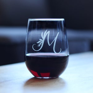 Könnte beinhalten: Ein klares, stielloses Weinglas aus Glas mit einem weißen, geätzten Blumendesign und dem Buchstaben "M" an der Seite. Das Glas ist mit Rotwein gefüllt.