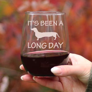 May include: A clear stemless wine glass with the text "It's been a long day" and a silhouette of a dachshund dog. The glass is filled with red wine.