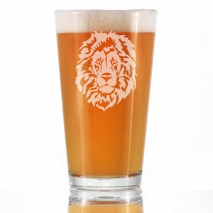May include: A clear glass pint glass with a white lion head design etched on the front. The glass is filled with a light amber colored beer.