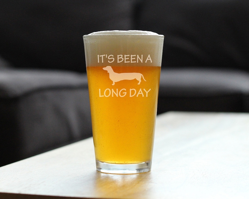May include: A clear glass pint glass with a white dachshund silhouette and the text "It's been a long day". The glass is filled with a golden yellow beer.