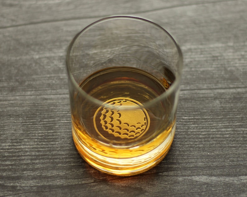 May include: A clear glass tumbler with a gold golf ball design etched into the bottom. The tumbler is filled with a golden liquid.