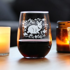May include: A stemless wine glass filled with red wine. The clear glass features a white etched design of a rabbit surrounded by floral elements. The glass is set on a wooden surface, with a lit candle and a jar in the background.