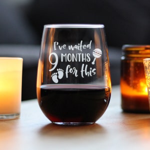May include: A clear glass wine glass with the text "I've waited 9 months for this" etched on the side. The glass is filled with red wine and there are small baby footprints and a rattle etched on the side.