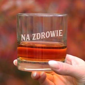 Puede incluir: Un vaso de vidrio transparente con un líquido marrón en su interior. El vaso tiene las palabras "NA ZDROWIE" grabadas en la parte delantera.