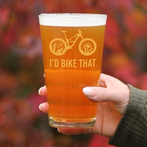 Op de afbeelding: Een helder glazen pintglas met een silhouet van een mountainbike en de tekst "I'd Bike That". Het glas is gevuld met een licht amberkleurig bier.