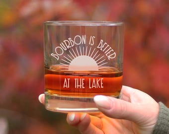 以下が含まれることがあります： 側面に「Bourbon is better at the lake」という文字がエッチングされた透明なガラス。ガラスは茶色の液体で満たされており、太陽のグラフィックと光線が描かれています。