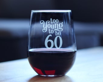 May include: A clear glass stemless wine glass with the text "too young to be 60" etched on the side. The glass is filled with red wine.
