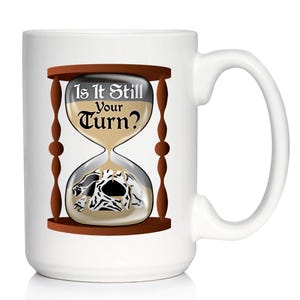 Pode incluir: Caneca de cerâmica branca com um design de ampulheta marrom. A ampulheta contém areia e uma caveira, com o texto "Is It Still Your Turn?" acima. A caneca tem uma alça curva.