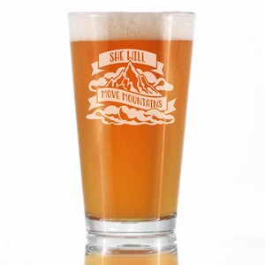 May include: A clear pint glass filled with a light amber liquid and topped with a foamy head. The glass features the etched phrase "She Will Move Mountains" with a mountain graphic and cloud accents.