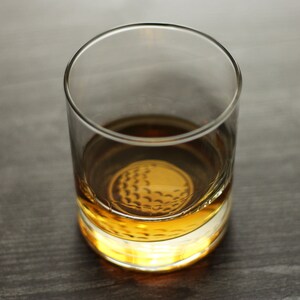 May include: A clear glass tumbler with a golf ball design on the bottom. The tumbler is filled with a golden brown liquid.