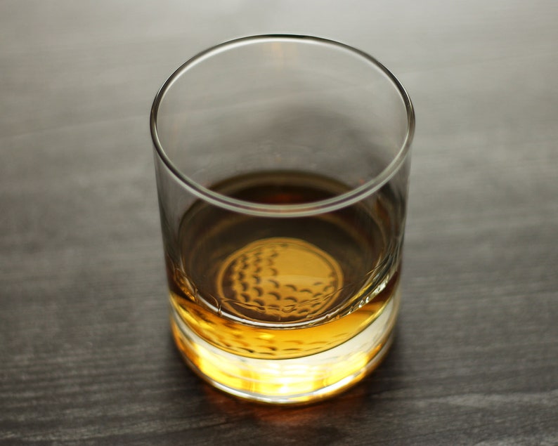 May include: A clear glass tumbler with a golf ball design on the bottom. The tumbler is filled with a golden brown liquid.
