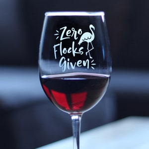 May include: A clear wine glass with a white flamingo and the text "Zero Flocks Given" etched on the side. The glass is filled with red wine.