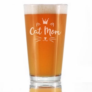 May include: A clear glass pint glass with a white design that says "Cat Mom" with a crown and cat whiskers. The glass is filled with a light amber colored beer.