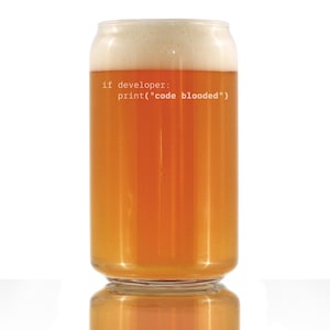 May include: A clear glass beer can-shaped glass filled with amber-coloured beer and a foamy white head. The text "if developer: print("code blooded")" is printed on the glass in a light grey colour. The glass is sitting on a white surface.