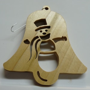 May include: A wooden Christmas ornament shaped like a bell with a snowman cutout. The snowman is wearing a hat and has a scarf. The ornament has a silver hanger.