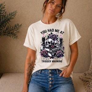 Può includere: T-shirt color crema con un design grafico con teschi, rose e candele. Il testo "YOU HAD ME AT TRIGGER WARNING" è stampato sotto la grafica. La modella indossa pantaloncini di jeans.